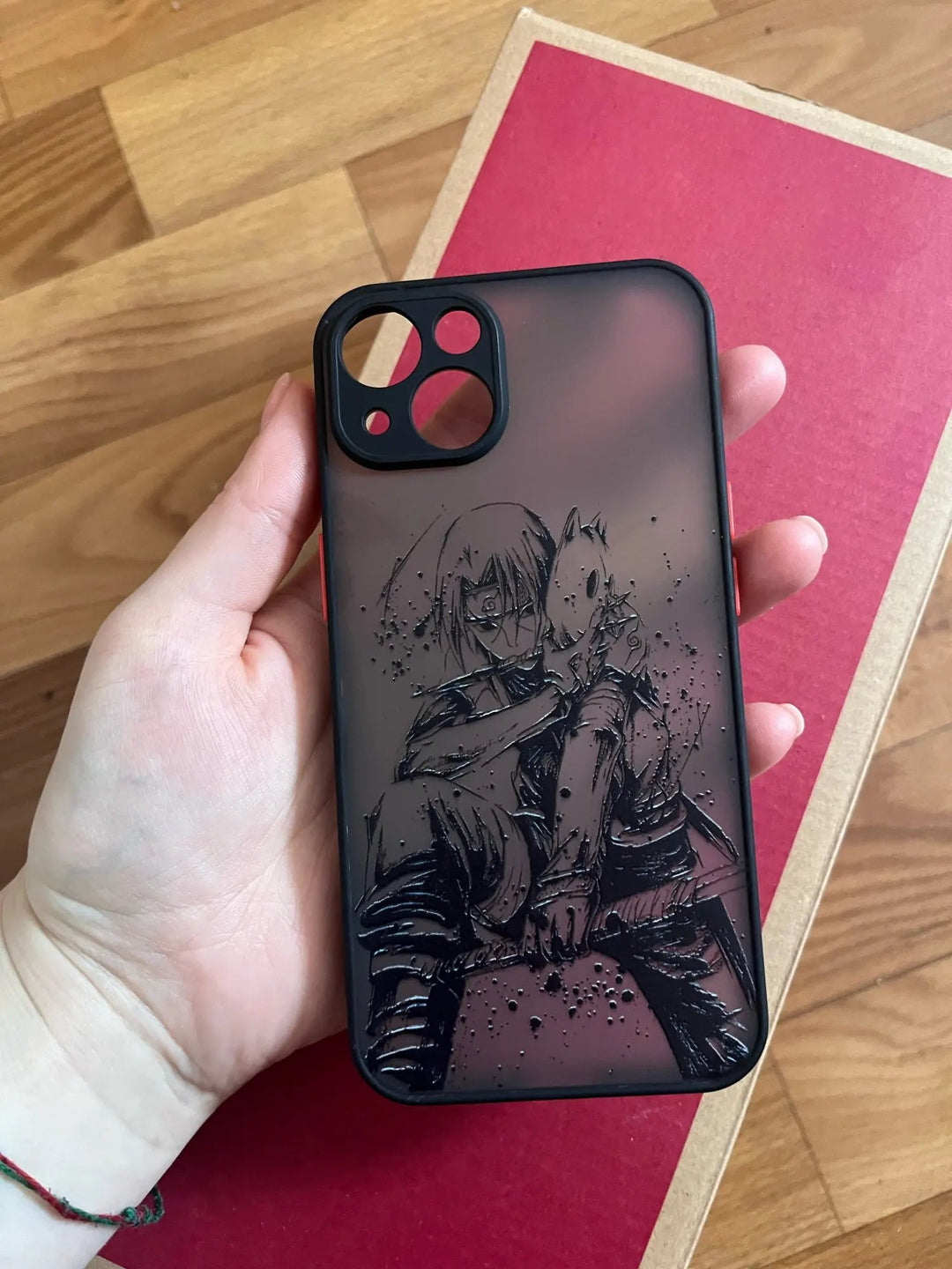 A hand holds the Anbu Translucent phone case, which features an illustrated character with spiky hair and a fox-like mask reminiscent of the ANBU, set against a red and wooden background.