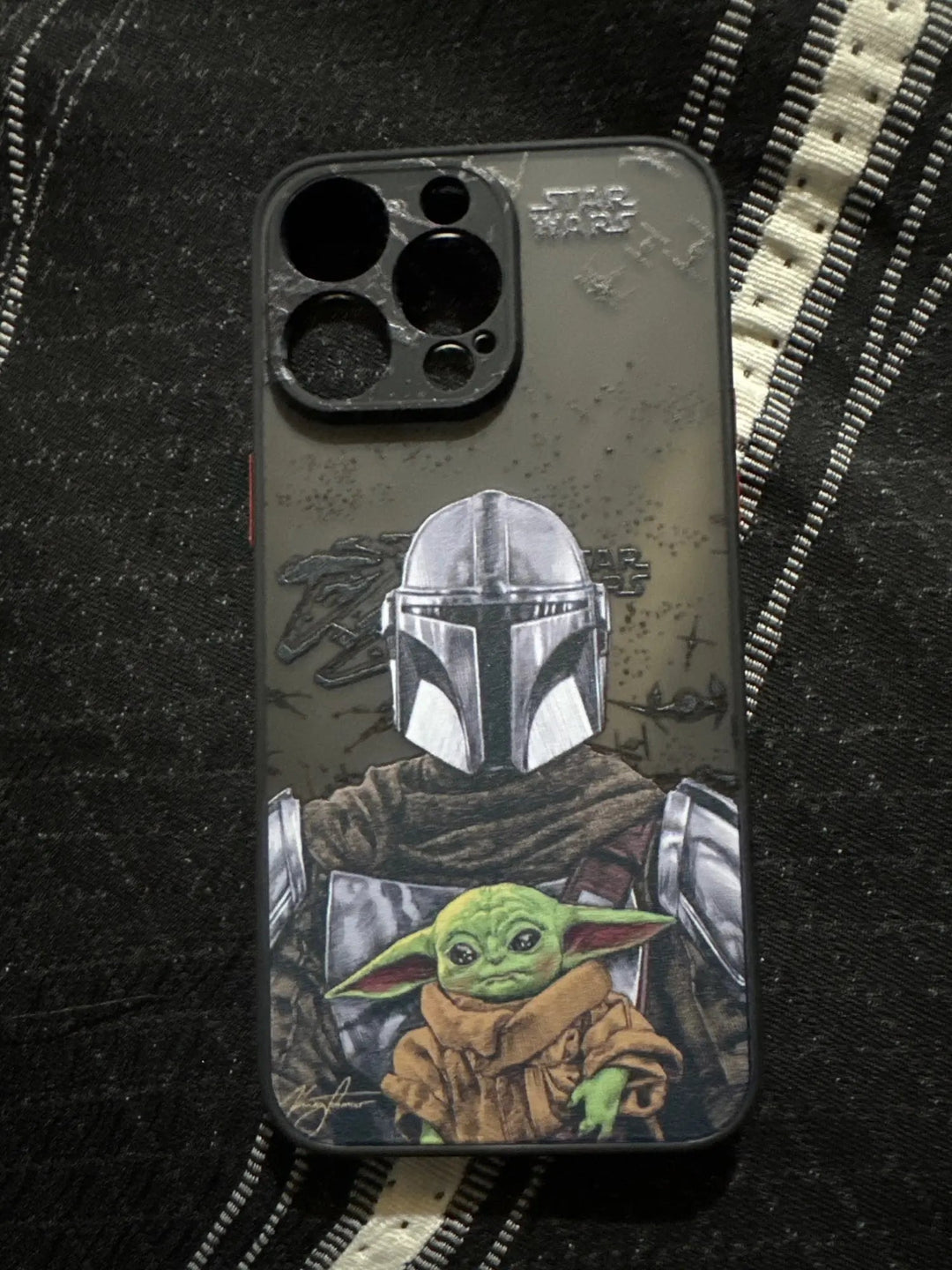 Phone case on a striped fabric background, featuring illustrations of a helmeted character and Baby Yoda from the well-known sci-fi series The Mandalorian, titled Bounty Hunter for iPhone.