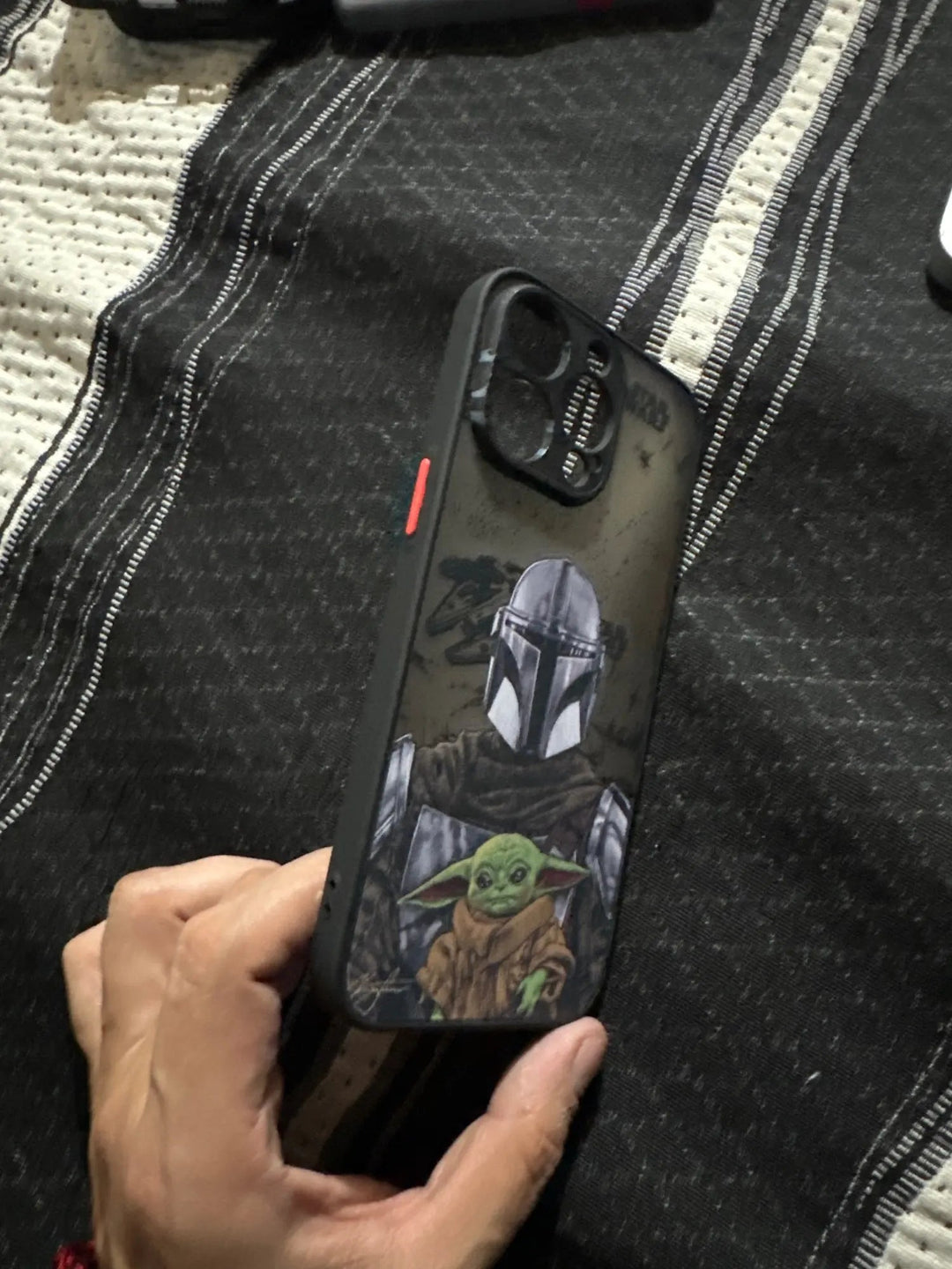 A hand holding an iPhone encased in the "Bounty Hunter" design, featuring The Mandalorian and Grogu, set against a striped blanket background.