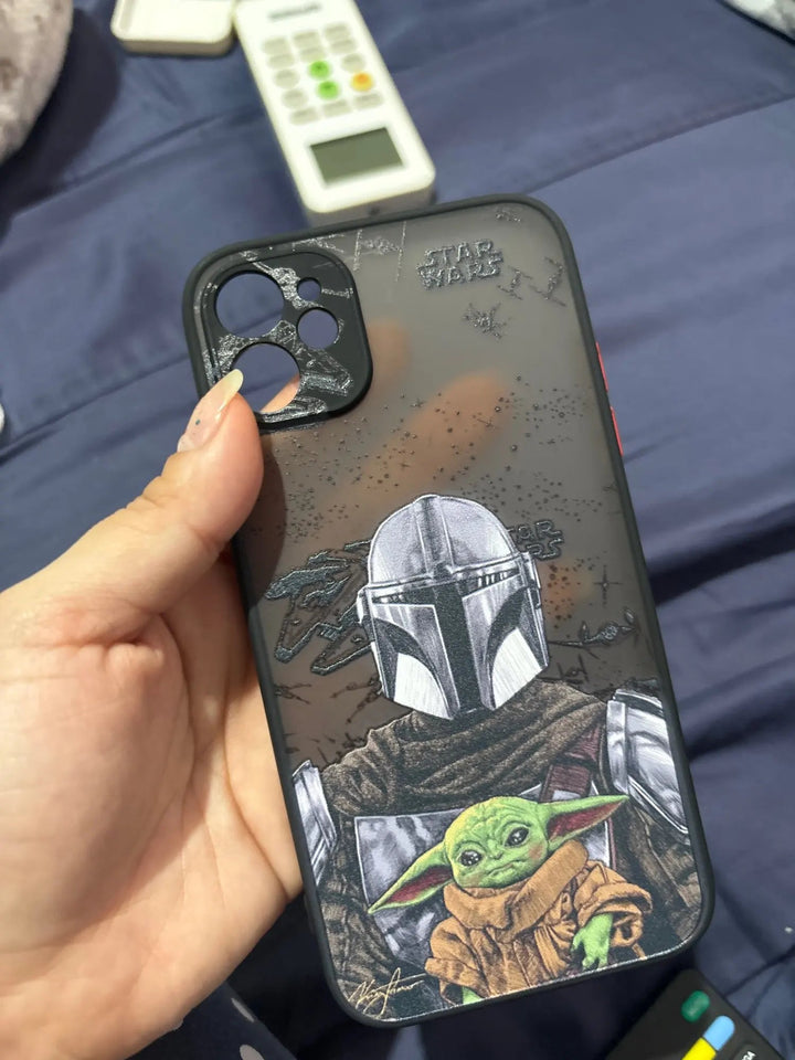 A person holds a Bounty Hunter for iPhone case featuring characters from The Mandalorian and Baby Yoda, with a cracked camera lens area. A remote control is visible in the background.