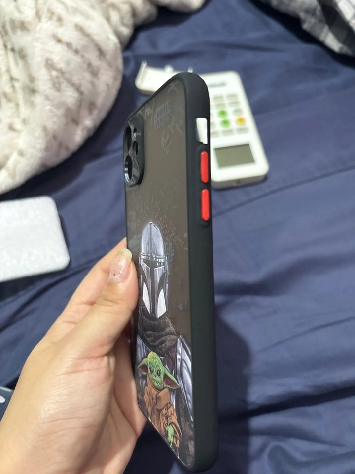 A hand holds a smartphone encased in the Bounty Hunter for iPhone case, adorned with characters from the Star Wars series, including Grogu from The Mandalorian, on a bed with a blanket and a calculator visible in the background.