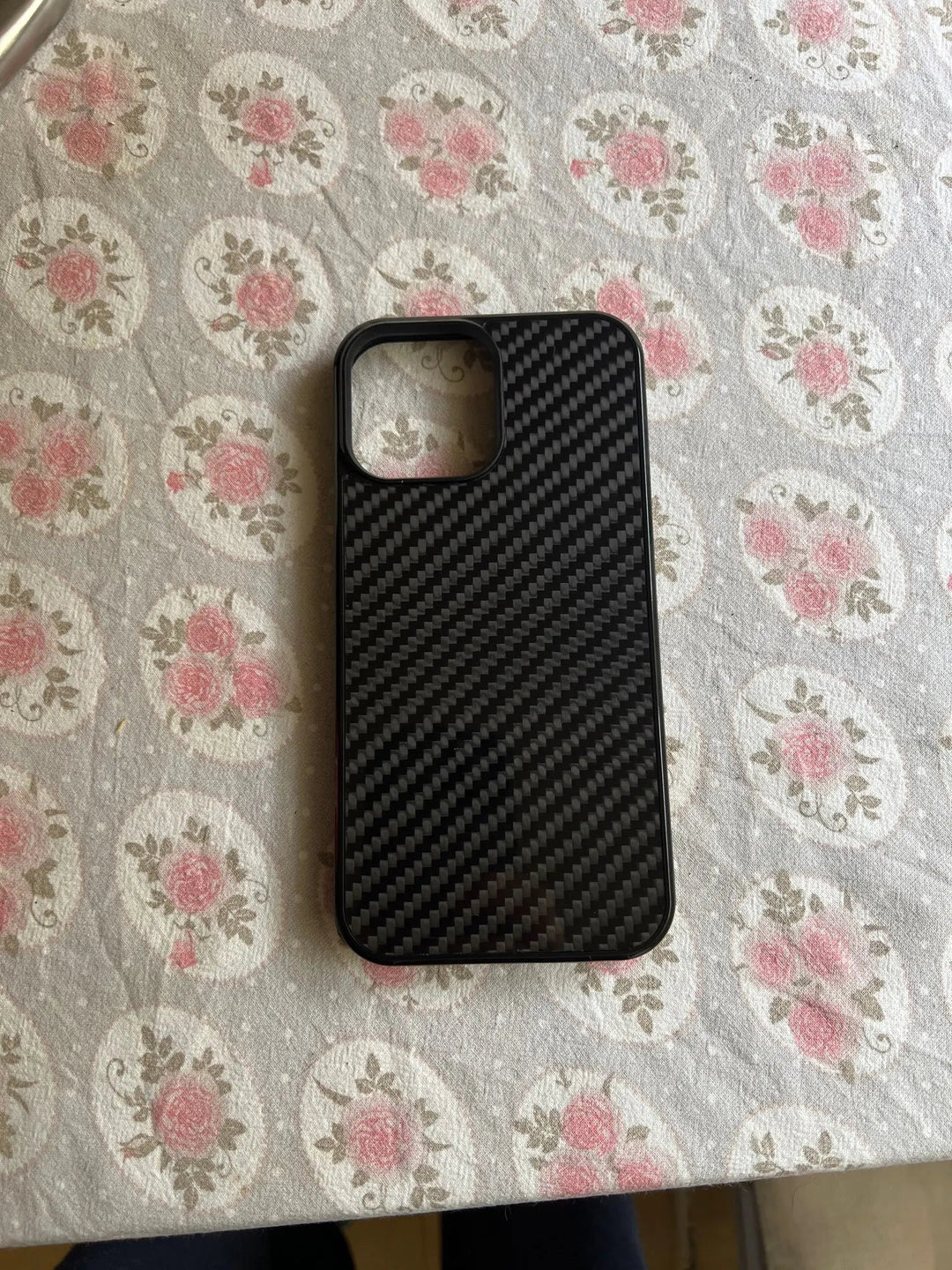 A sleek Carbon Fiber for iPhone lies on a white and pink floral-patterned tablecloth.