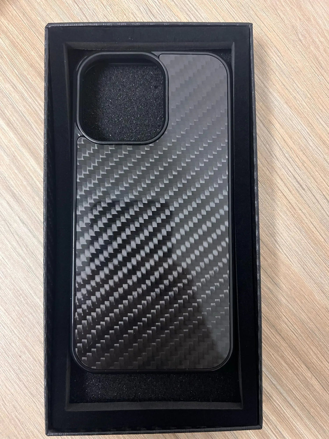 A sleek Carbon Fiber for iPhone case is placed inside a black rectangular box on a wooden surface.