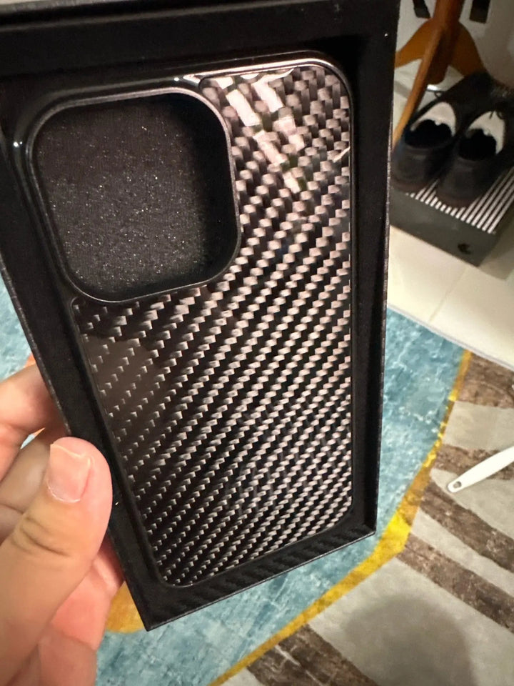 Hand holding a Carbon Fiber for iPhone phone case in packaging. A rug and a pair of shoes are visible in the background.