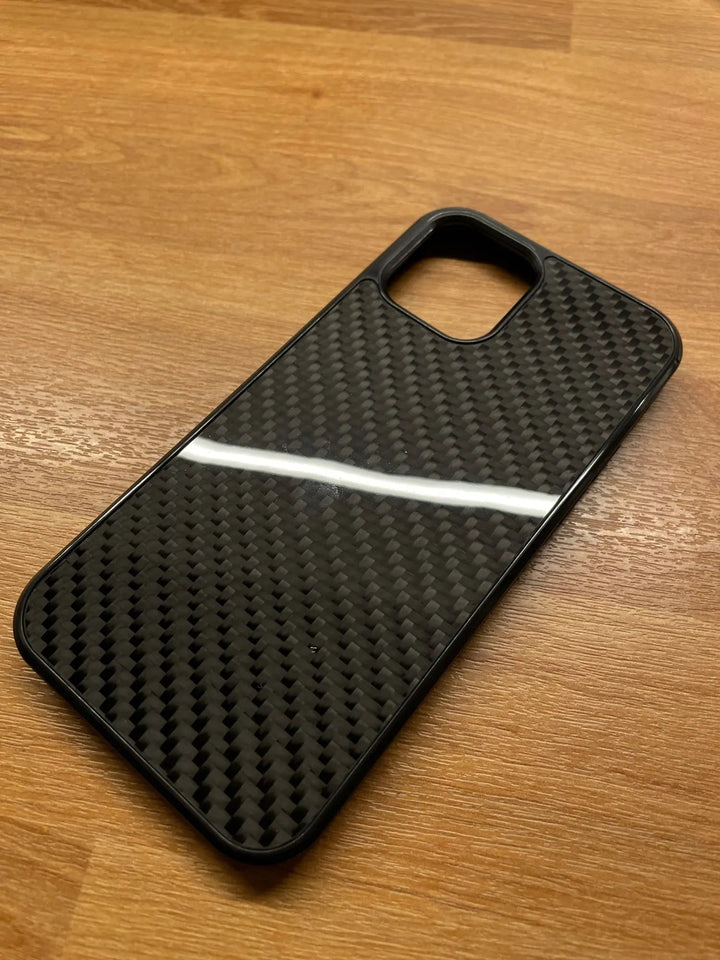 A sleek Carbon Fiber for iPhone, made from Aramid carbon fiber, is lying on a wooden surface. The black case, designed for optimal iPhone protection, features a rectangular opening for the camera.