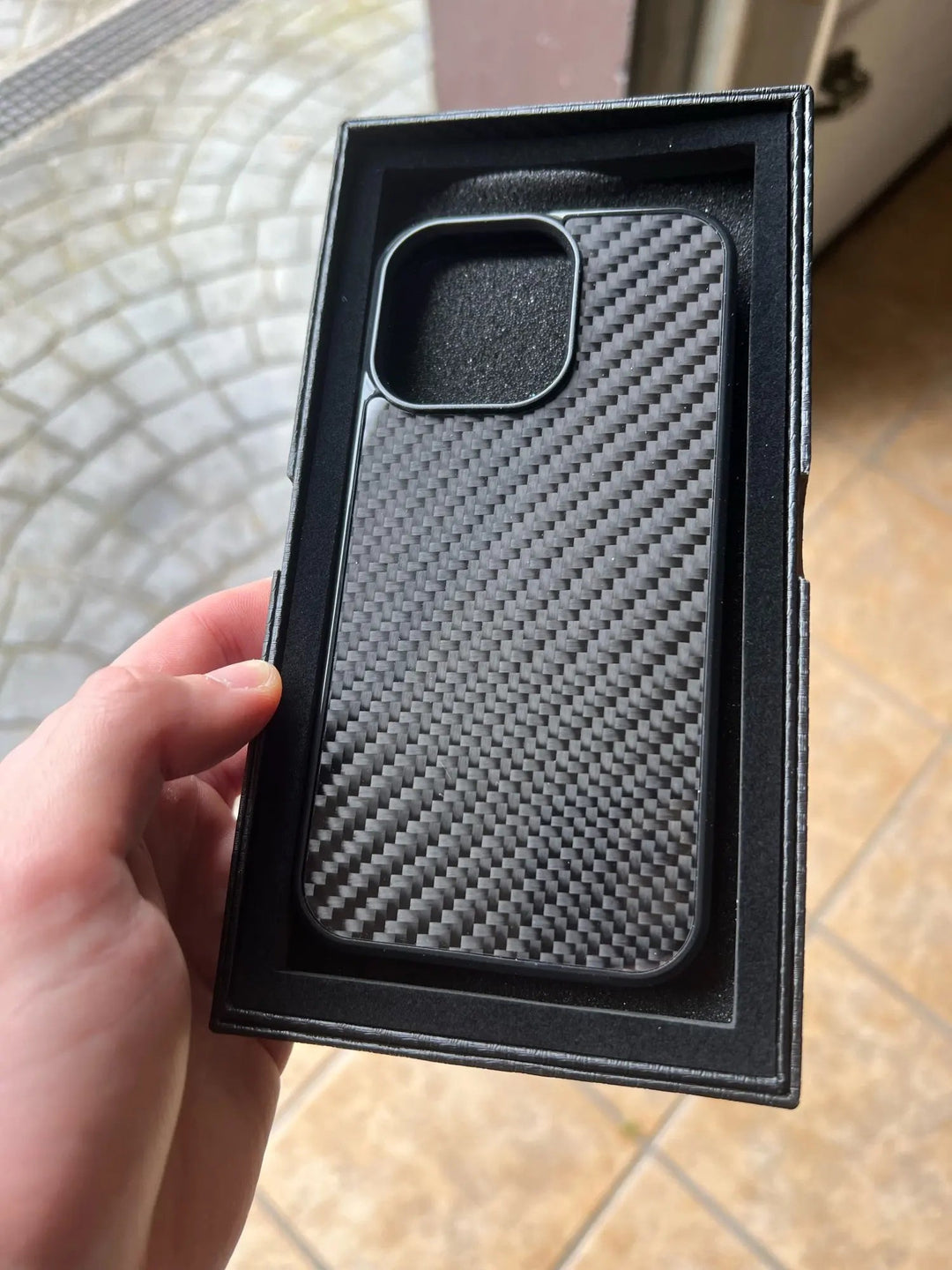 A hand holds a sleek Carbon Fiber for iPhone case inside its packaging, offering superior protection.