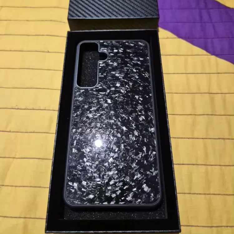 The Carbon Fiber for Samsung phone case, featuring a sleek black and white marbled design, is displayed inside an open black box with a textured lid. The case's glossy surface reflects light, ensuring optimal protection for Samsung devices. The box lies on a yellow and purple quilted background.