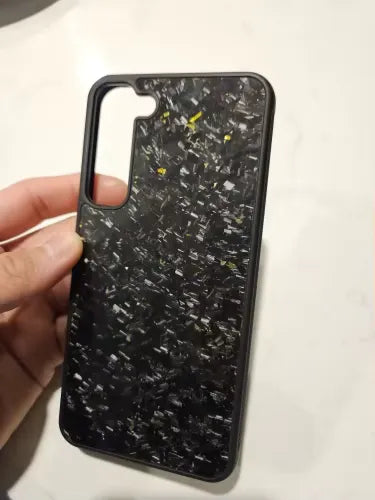 A hand is holding the Carbon Fiber for Samsung, a black phone case featuring a speckled texture. The case has a large cutout for the camera module and appears to have a glossy finish with small white and yellow specks scattered across its surface, designed to offer ultimate protection for Samsung devices.