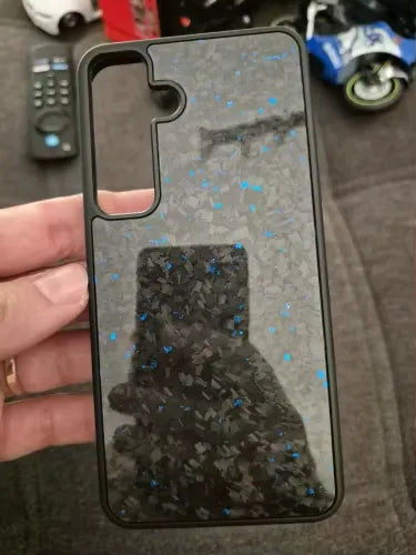 A hand holding the Carbon Fiber for Samsung smartphone case, which features a glittery black back adorned with blue and silver specks. The reflective surface of the case captures a partial reflection of the hand and a remote control in the background, offering ultimate protection for Samsung devices.