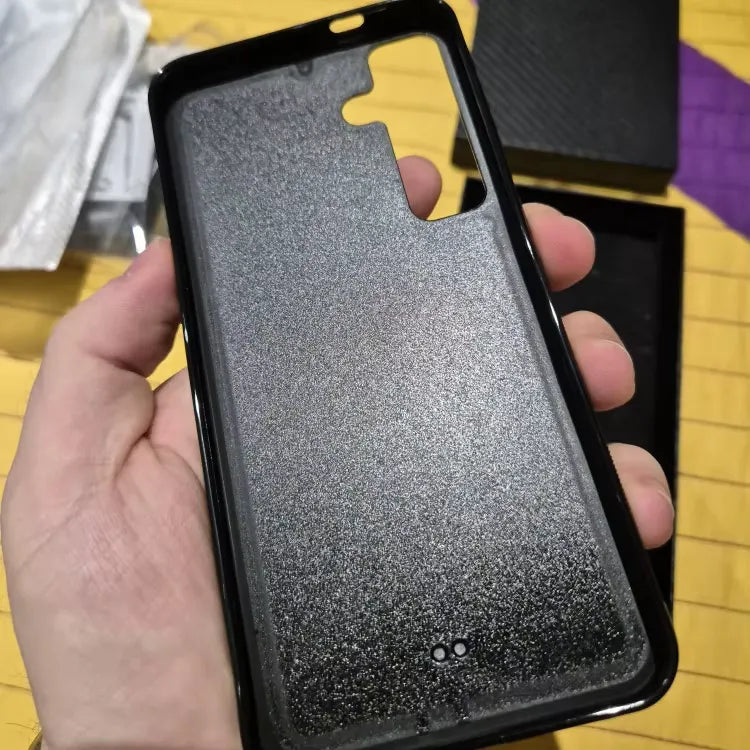 A hand holding a Carbon Fiber for Samsung case with a matte, glittery texture. The case ensures ultimate protection with precise cutouts for the camera, flash, and other ports. There is a box in the background along with some other indistinguishable items on a yellow surface.