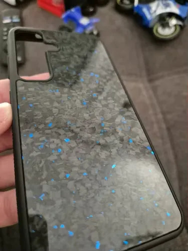 A close-up view of the Carbon Fiber for Samsung case, featuring a black and blue speckled pattern. The case is being held by a hand, offering ultimate protection for Samsung devices. Some blurred objects, including toy vehicles, are visible in the background.