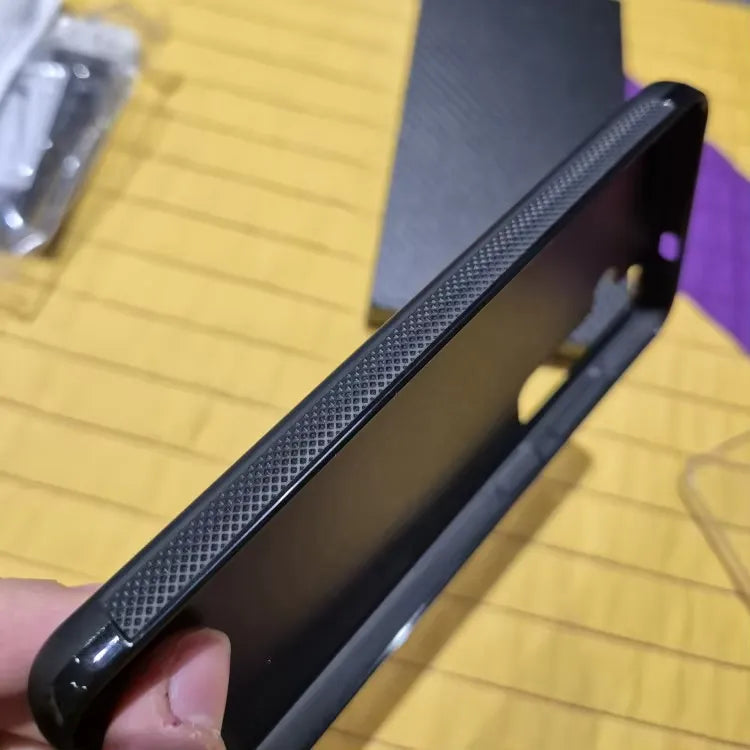 Close-up of a person holding the Carbon Fiber for Samsung, a black smartphone case with a textured grip on its side. The background features a wooden surface and some blurry objects, possibly phone accessories, laying around. This case offers ultimate protection and is compatible with Samsung devices.