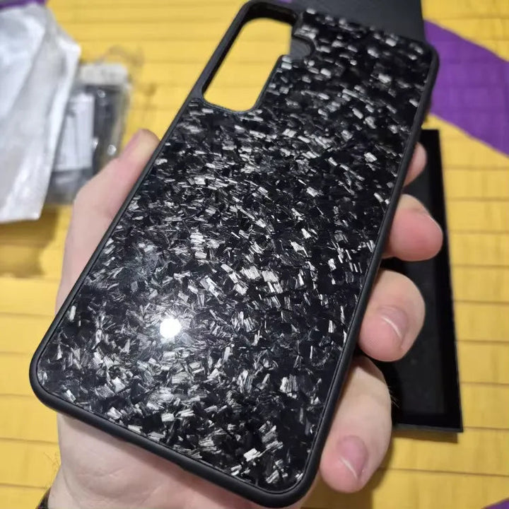 A hand holds the Carbon Fiber for Samsung smartphone case, which has a glossy, textured pattern featuring numerous small, reflective shards. The black case offers ultimate protection and has a large camera cutout. The person is indoors with various items and a yellow and purple surface in the background.