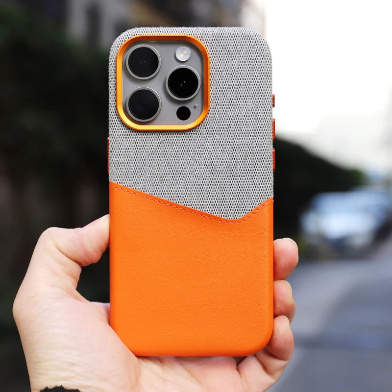 A hand holding a smartphone with a card holder case, featuring a gray and orange two-toned design, three camera lenses on the back, and MagSafe compatibility. The background is blurred.