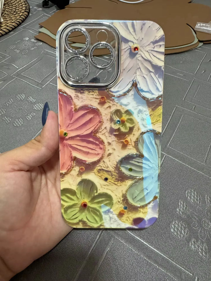 A photo of a phone case with a 3D floral design. The flowers are painted in pastel colors and adorned with glitter and rhinestones. The case is being held in someone's hand.