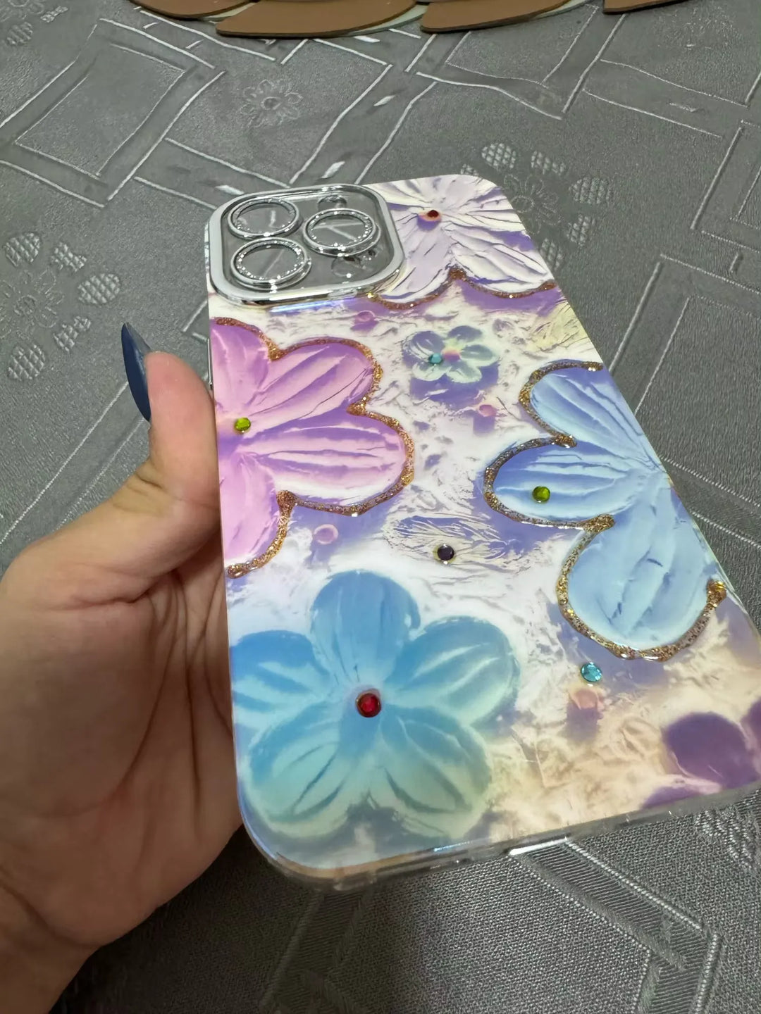 A photo of a phone case with a 3D floral design. The flowers are painted in pastel colors and adorned with glitter and rhinestones. The case is being held in someone's hand.