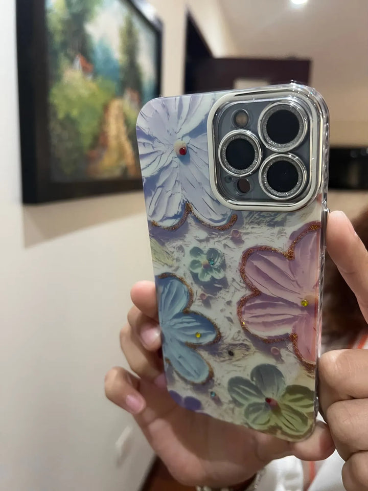 A photo of a phone case with a 3D floral design. The flowers are painted in pastel colors and adorned with glitter and rhinestones. The case is being held in someone's hand.