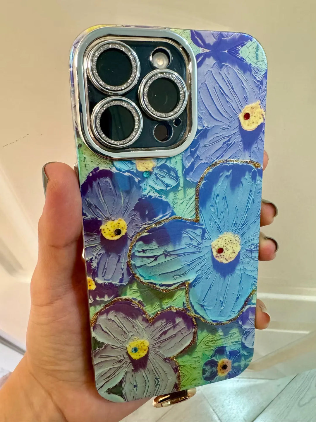 A photo of a phone case with a 3D floral design. The flowers are painted in pastel colors and adorned with glitter and rhinestones. The case is being held in someone's hand.