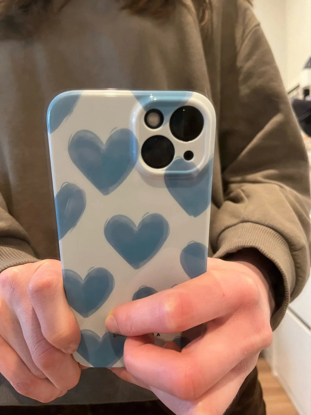 A person holding an iPhone encased in the Couple Hearts for iPhone - Acrylic case, featuring a slim, playful heart pattern in white and blue, faces the camera while wearing a brown sweater.