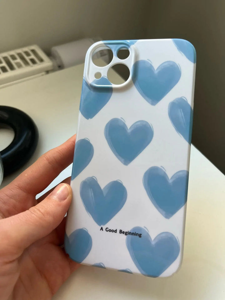 A hand holds an iPhone encased in a slim, acrylic case with playful blue hearts and the text "A Good Beginning" at the bottom.