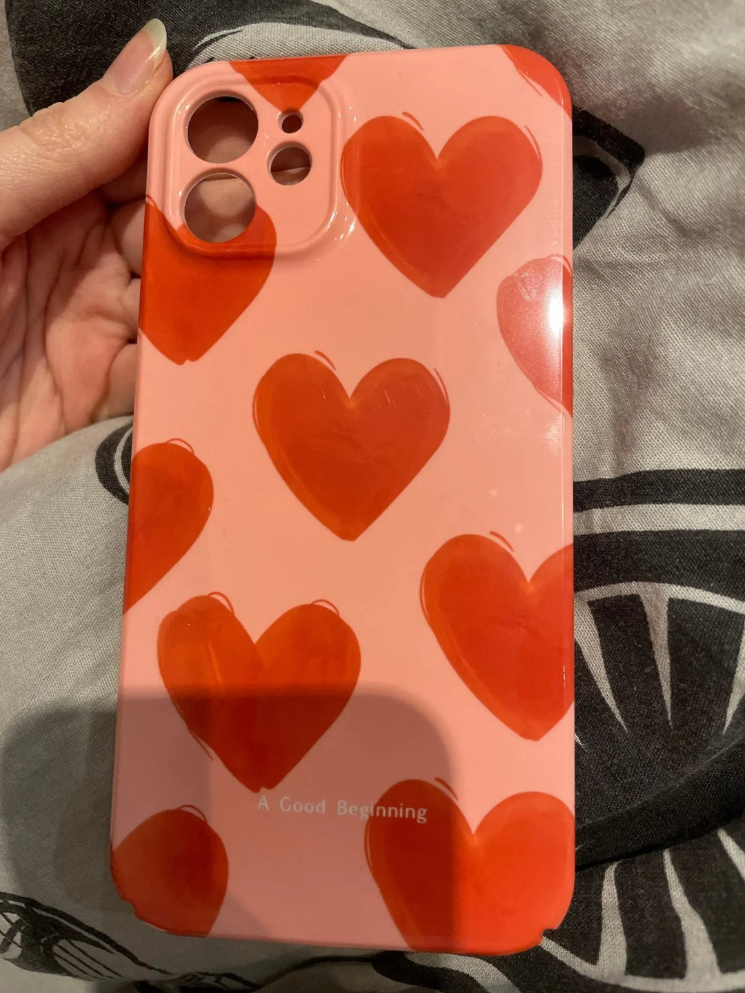 A hand holds the Couple Hearts for iPhone case, featuring a playful design with large red heart patterns on a pink background and text reading "A Good Beginning.