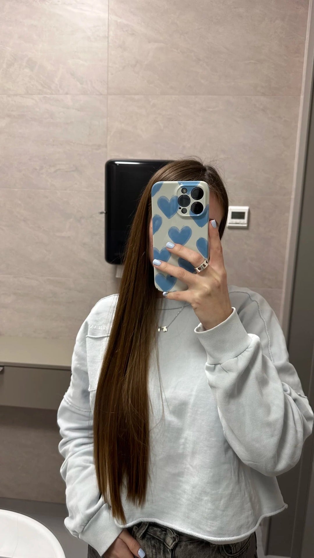 A person with long hair takes a mirror selfie in a bathroom, covering their face with an acrylic Couple Hearts case for iPhone that features playful heart patterns. They are wearing a light grey sweatshirt.