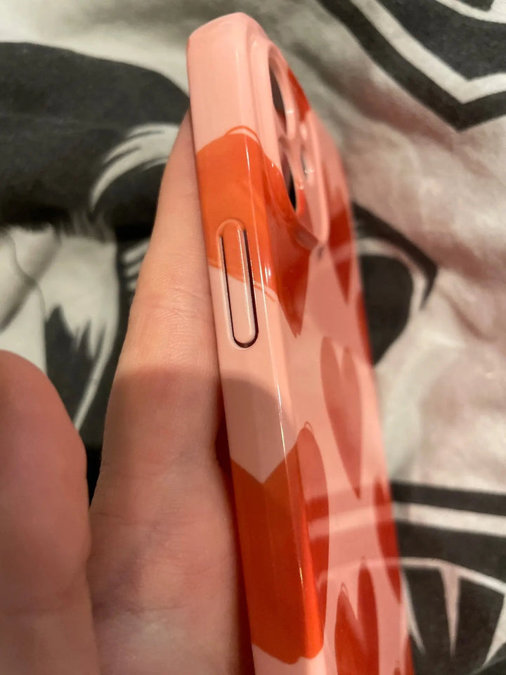 A close-up of a hand holding an iPhone adorned with the "Couple Hearts for iPhone - Acrylic" case, featuring a charming red and pink heart pattern. The phone's volume button is visible on the side, showcasing its playful design.