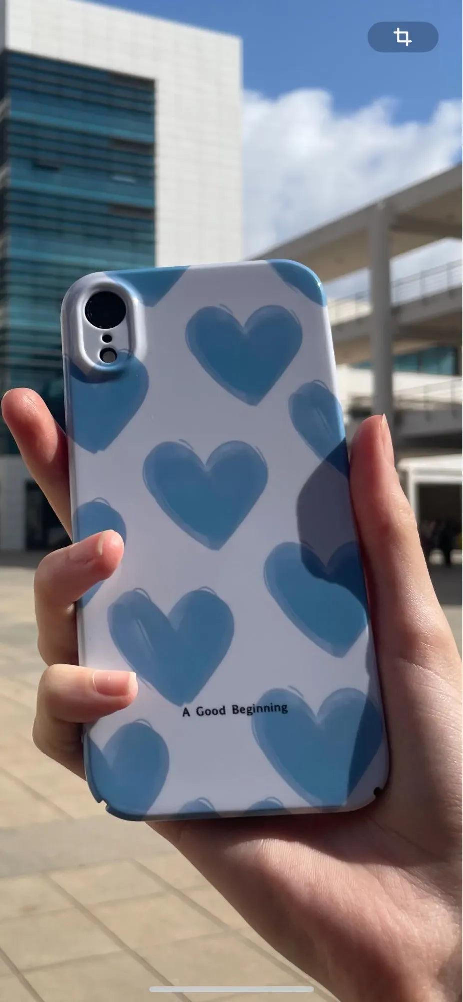 A hand holds a smartphone adorned with the Couple Hearts for iPhone - Acrylic case, showcasing a whimsical white and blue heart pattern with the phrase "A Good Beginning." A building is blurred in the background.