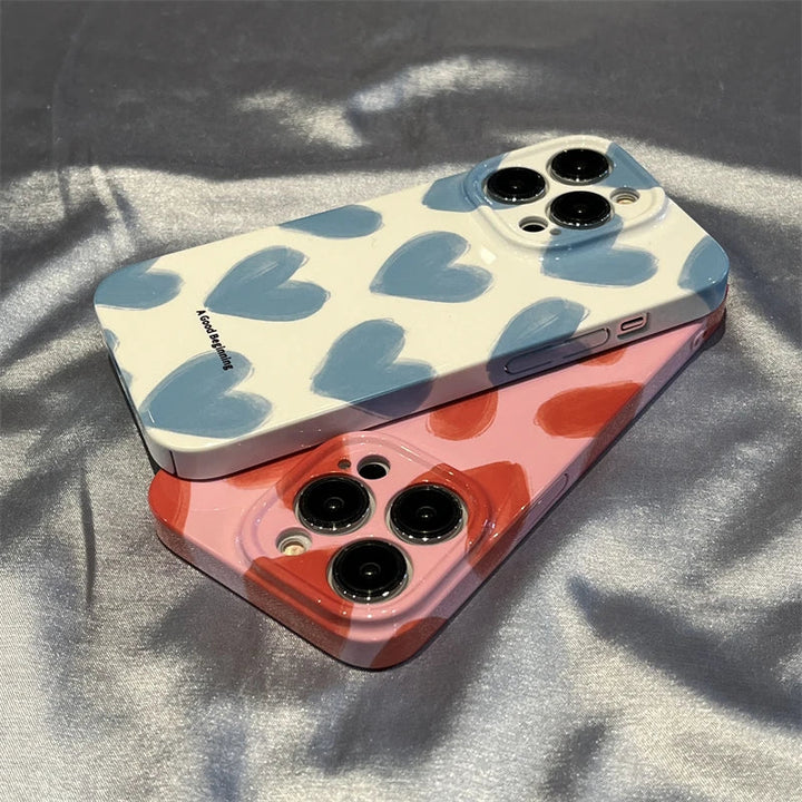 Two iPhones with Couple Hearts acrylic cases—one adorned in white with blue hearts and the other in pink with red hearts—are placed on a reflective surface, showcasing their playful design.