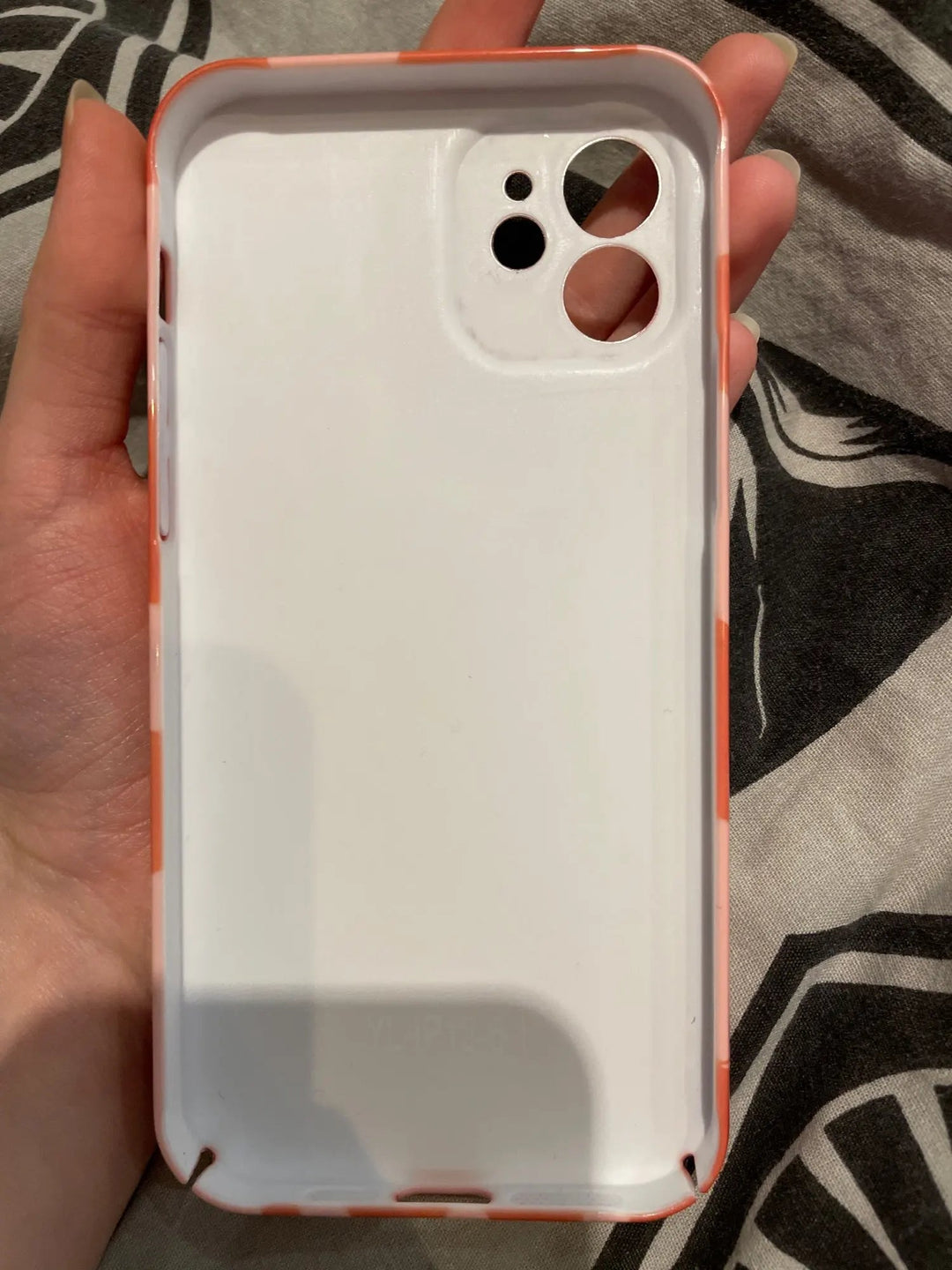 A hand holds the "Couple Hearts for iPhone" case, made of sleek white acrylic with an orange-trimmed edge and playful design cutouts for the camera and flash, all resting on a patterned fabric background.