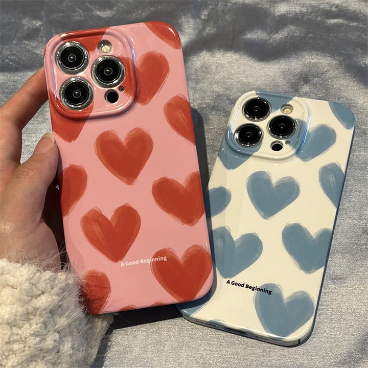 A hand holds a smartphone with the "Couple Hearts for iPhone - Acrylic" case featuring a pink heart pattern design, next to another phone with the same product line in a white case adorned with blue hearts. Both slim cases are part of the "A Good Beginning" collection and proudly display triple camera setups, offering a playful and charming design.