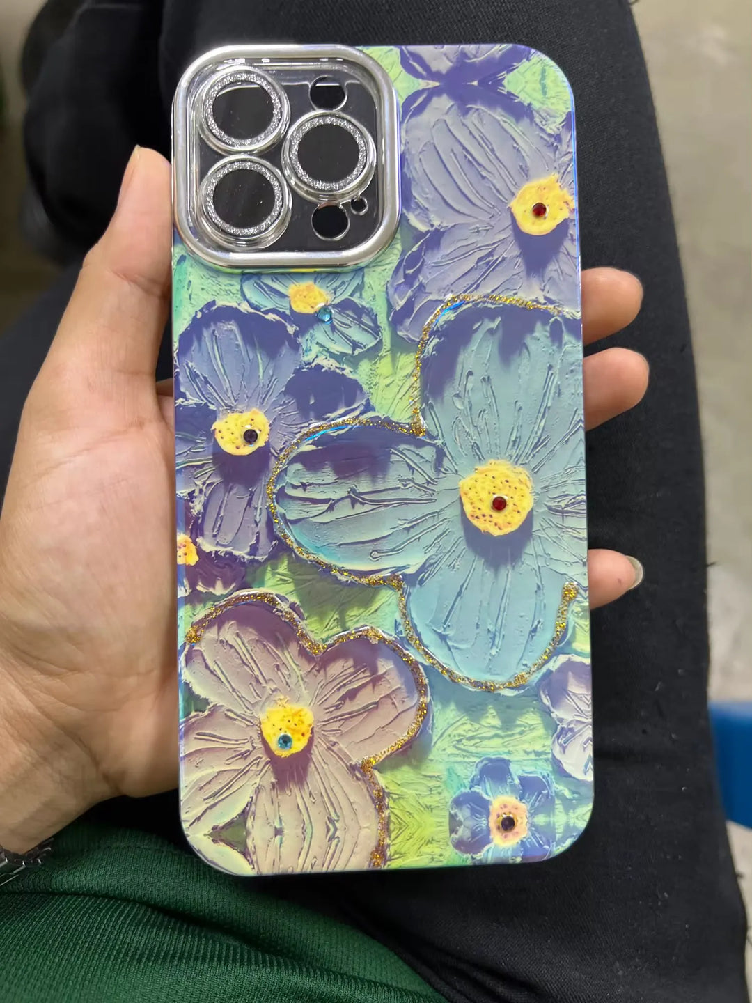 A close-up photo of a clear phone case with a 3D floral design. The flowers are painted in pastel colors and adorned with glitter and rhinestones. The case is being held in someone's hand.