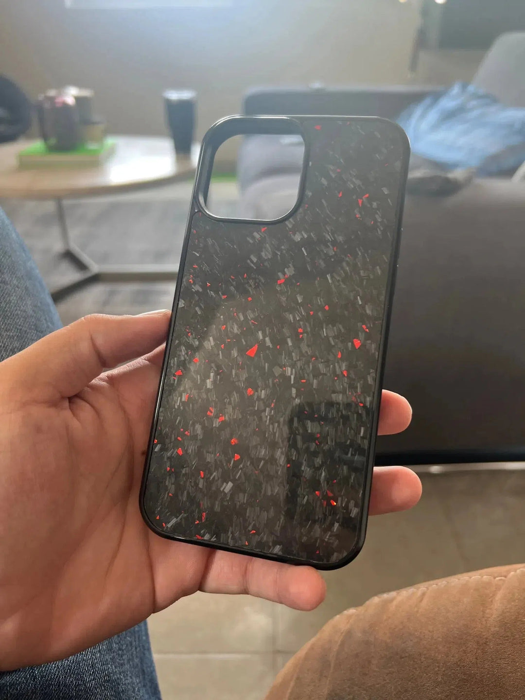 A person holds the Forged Carbon Fiber for iPhone - MagSafe case, featuring a speckled red and gray pattern on black, against a background showcasing a coffee table and couch.