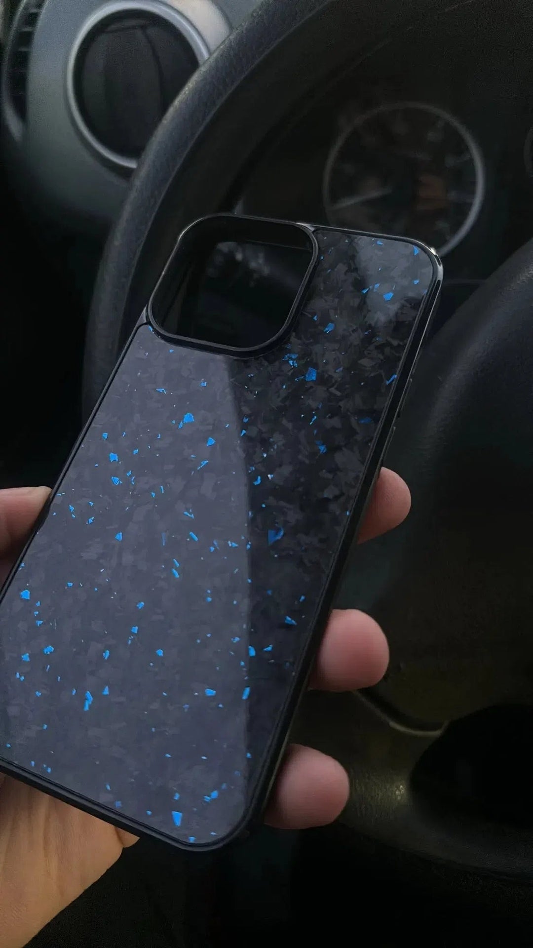 A hand holding the Forged Carbon Fiber for iPhone - Magsafe, a dark-colored phone case with scattered blue speckles, designed with sleek aramid carbon fiber, in front of a car steering wheel and dashboard.