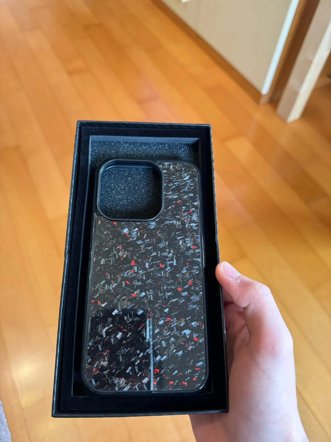 A hand is holding the Forged Carbon Fiber for iPhone - Magsafe, a black phone case with a speckled pattern of red and blue colors made from aramid carbon fiber, inside a rectangular black box. The background features a wooden floor.