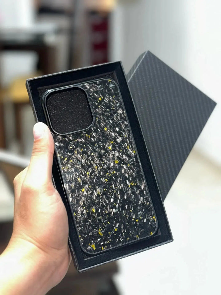 A hand holds a Forged Carbon Fiber for iPhone - Magsafe phone case, featuring a black and yellow speckled design, inside an open black box with a patterned lid, showcasing its sleek Aramid carbon fiber construction.