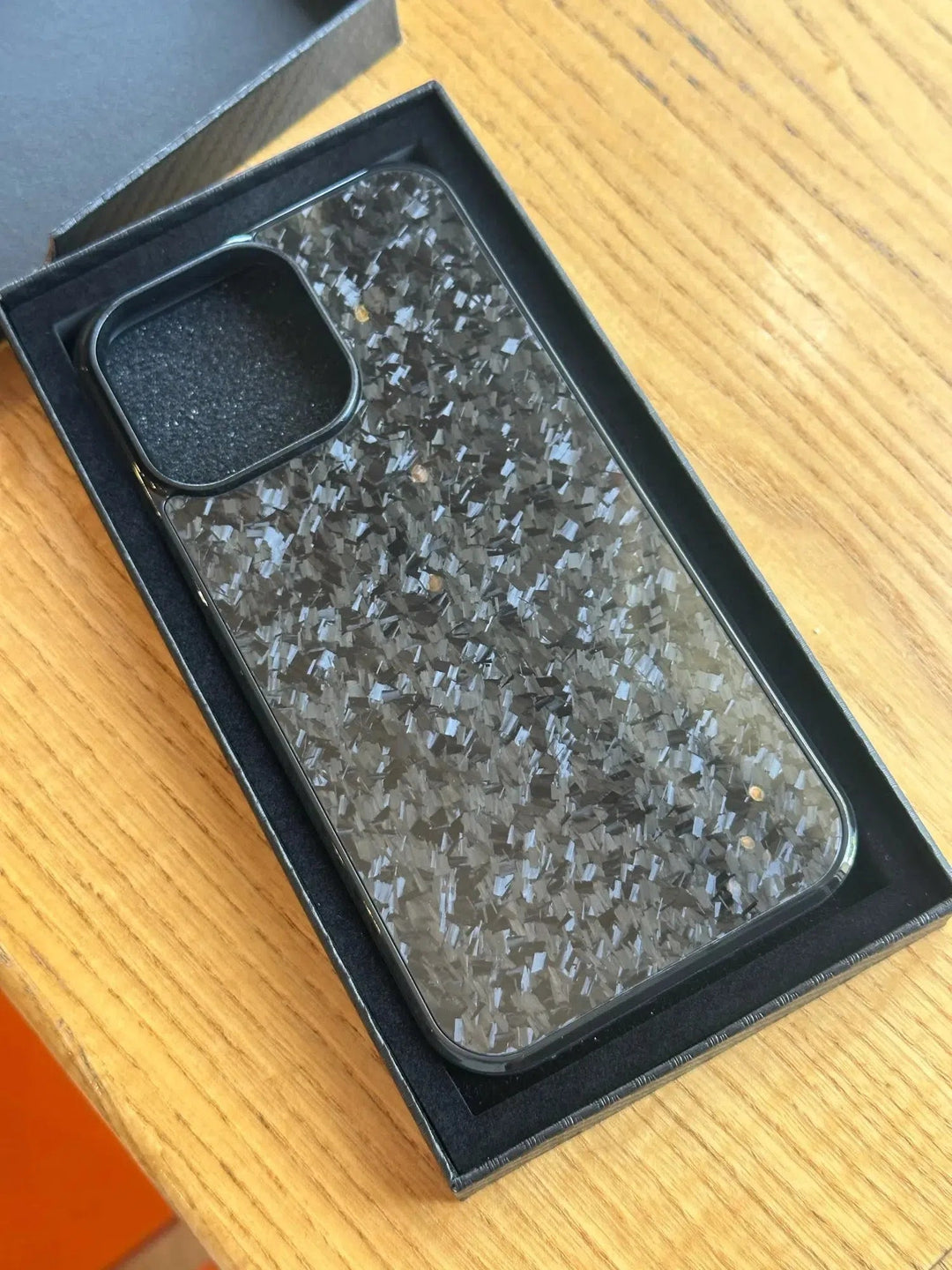 A Forged Carbon Fiber for iPhone - Magsafe case, featuring a textured, speckled appearance in black, is placed inside a black box on a wooden surface.