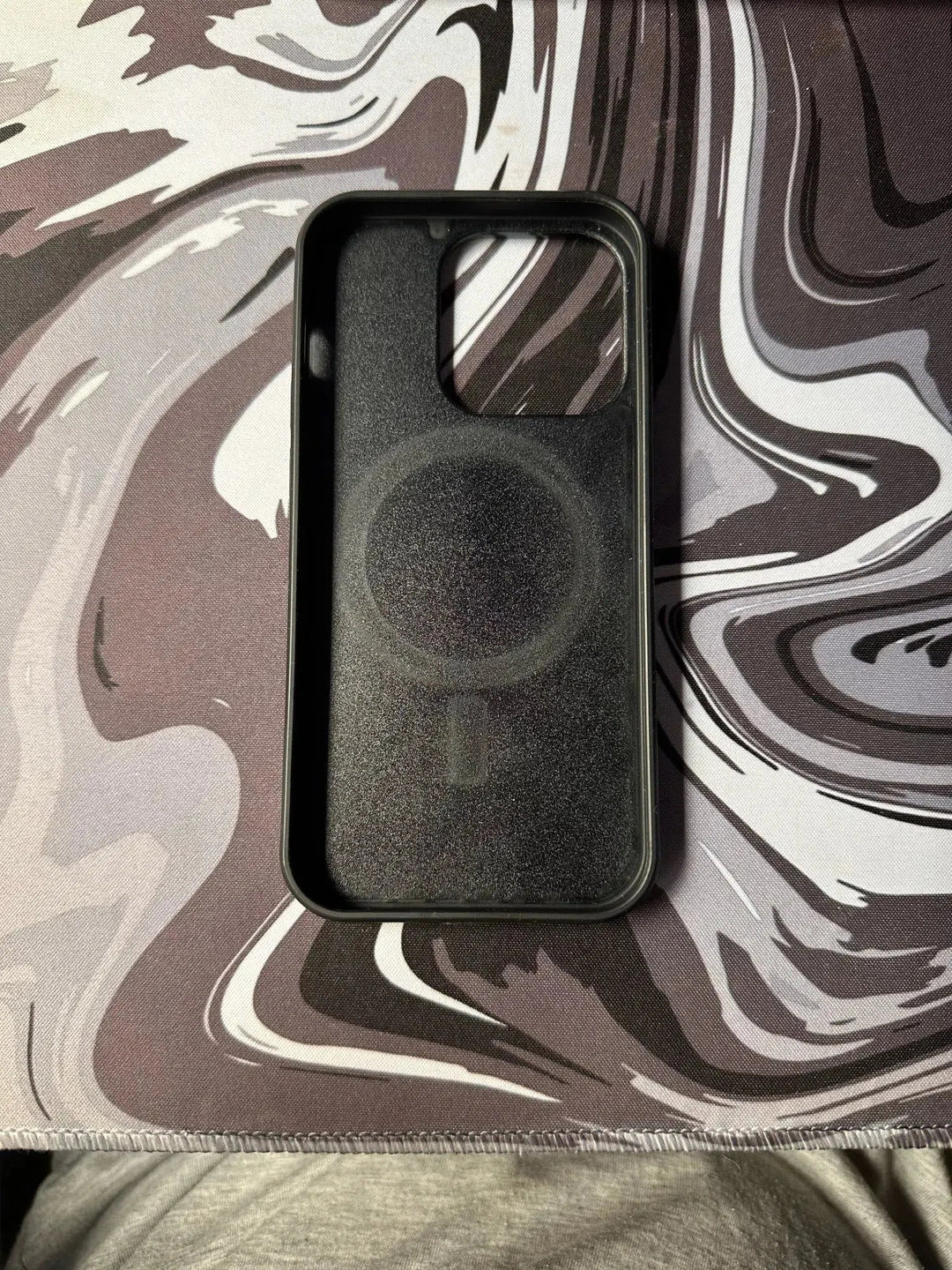 A Forged Carbon Fiber for iPhone - Magsafe case, featuring a cutout for the camera and a charging symbol on the inside, placed on a marbled surface background.