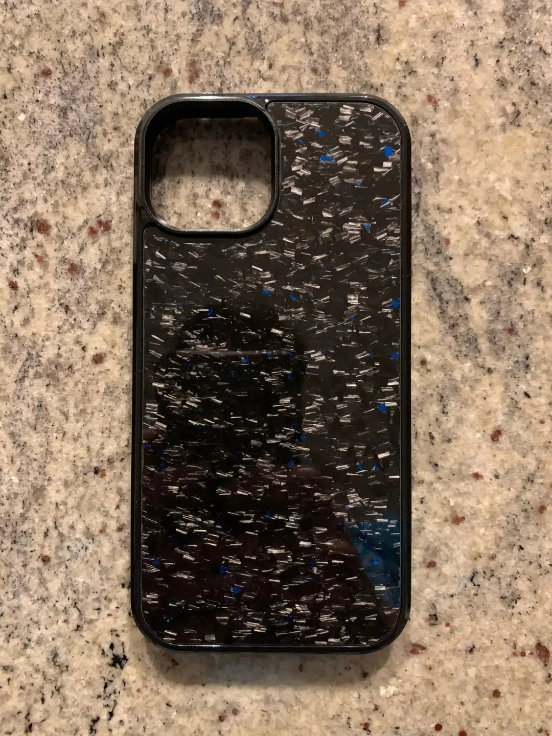 A Forged Carbon Fiber for iPhone - MagSafe with a speckled pattern of silver and blue, resting on a granite countertop.