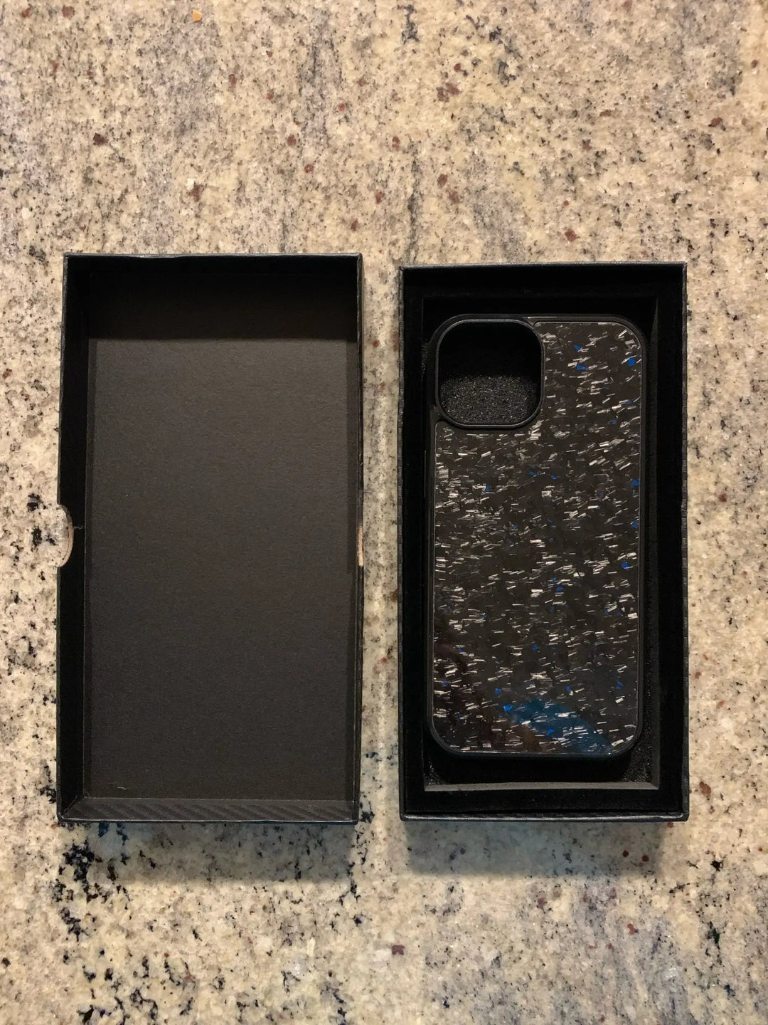 A black and blue Forged Carbon Fiber for iPhone - Non Magsafe case with a speckled design is placed inside a black open box. An empty black box sits beside it on a speckled countertop, showcasing the ultra-thin lightweight build made from aramid carbon fiber.