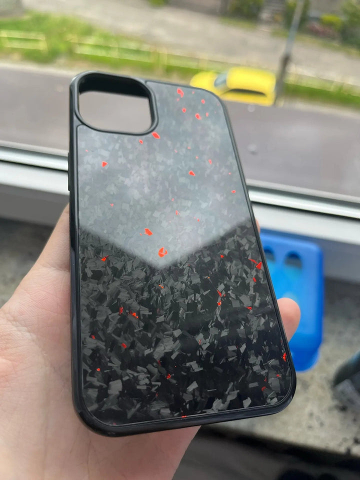 A hand holds the Forged Carbon Fiber for iPhone - Non Magsafe case, featuring a sleek black design with a glossy finish and scattered red and gray speckles, crafted from aramid carbon fiber. The background reveals a window with a blurry view of a road and a yellow car.