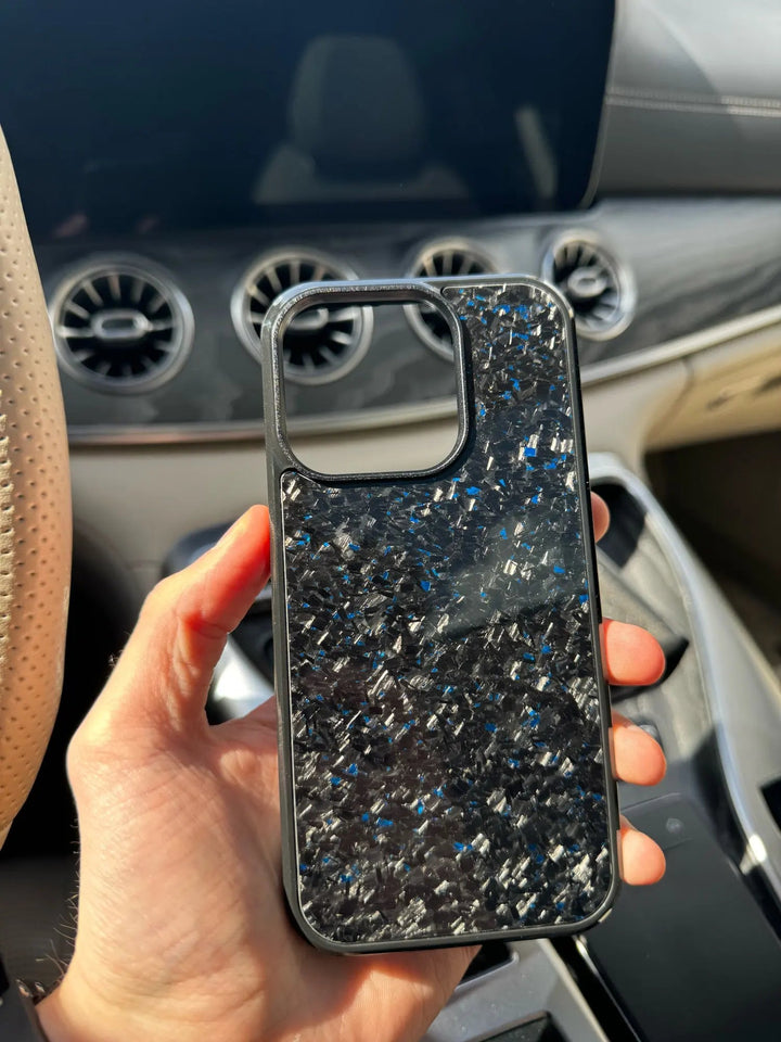 A hand holds a Forged Carbon Fiber for iPhone - Non Magsafe phone case inside a car. The interior of the car, including the dashboard and steering wheel, is visible in the background. The ultra-thin lightweight design of the case adds to its sleek appearance, seamlessly blending functionality with style.