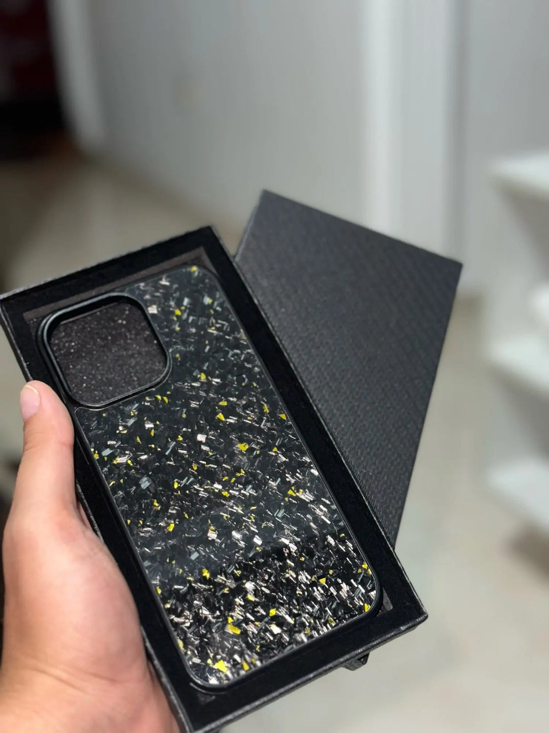 A hand holding an ultra-thin, lightweight Forged Carbon Fiber iPhone case with a speckled design made of aramid carbon fiber, displayed in a black box with an open lid.