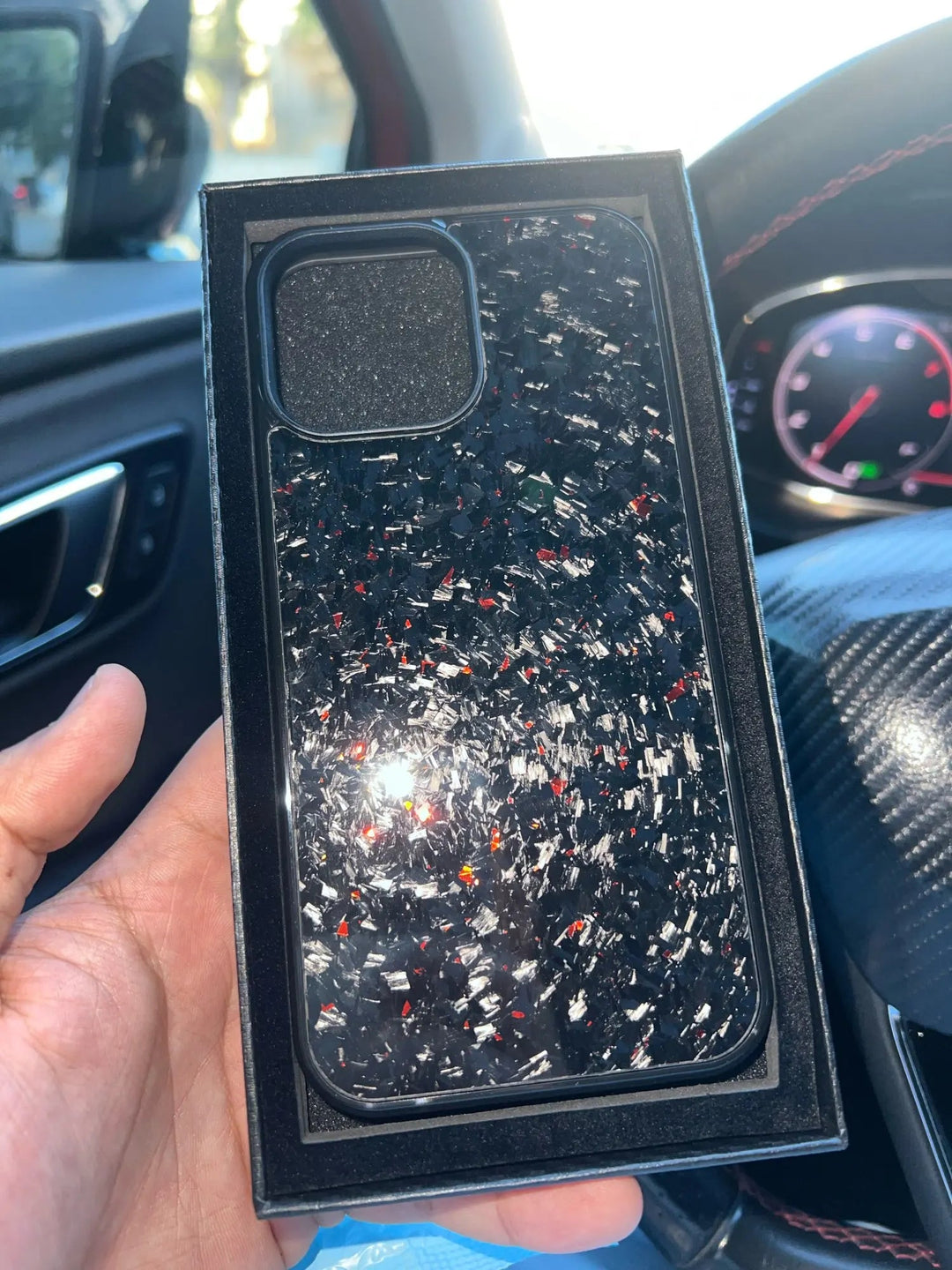 A hand is holding the Forged Carbon Fiber for iPhone - Non Magsafe, a sleek, ultra-thin lightweight black speckled phone case inside a box, with a car interior visible in the background.