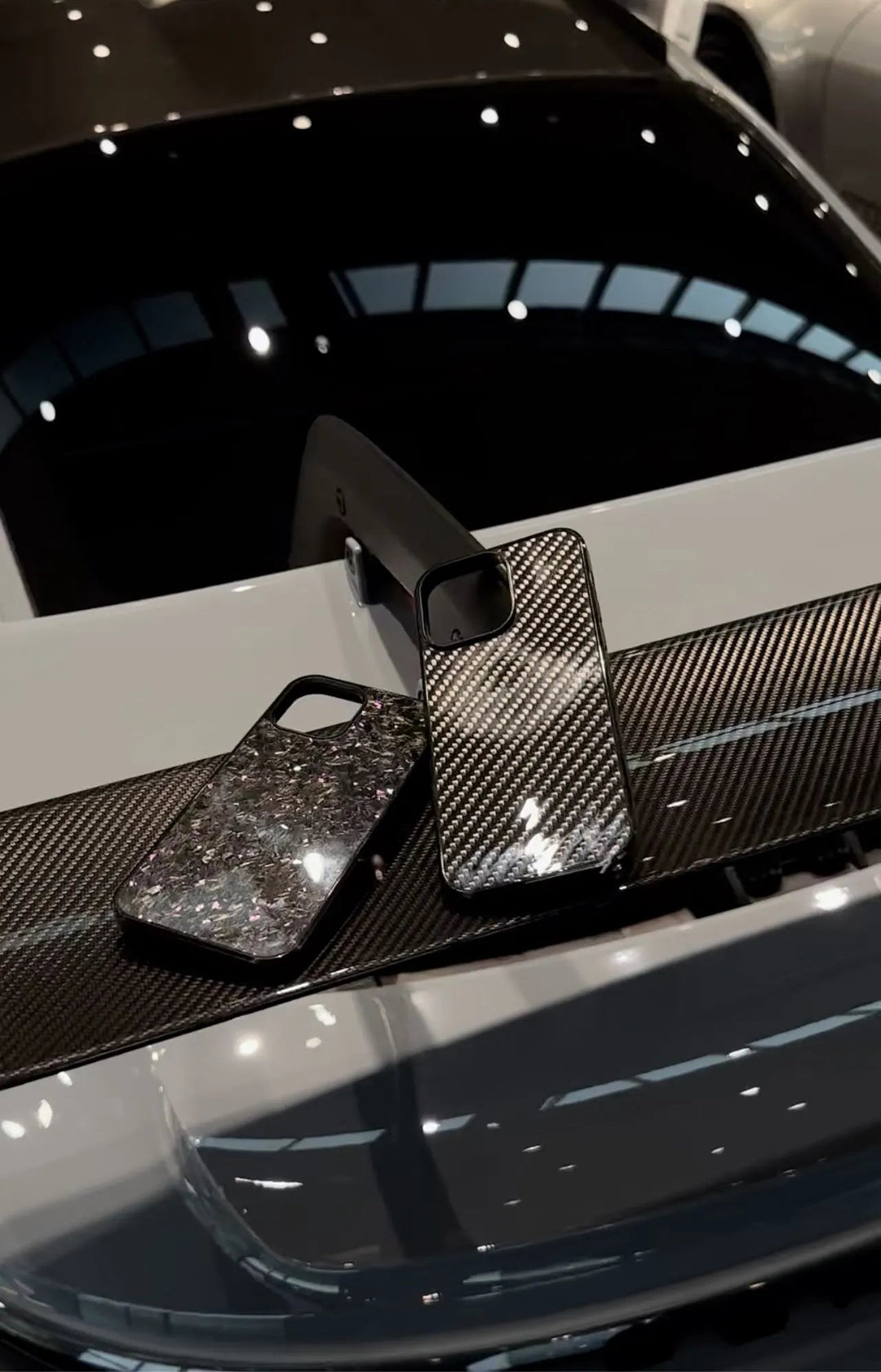 Two smartphone cases rest on the spoiler of a car, one with a glittery design, the other being the Forged Carbon Fiber for iPhone - Non Magsafe with an ultra-thin lightweight carbon fiber pattern. The car's rear window and part of its body are visible in the background.