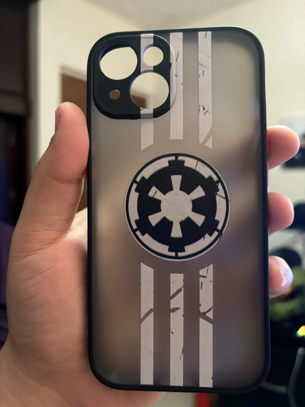 A hand holds a Galactic Empire Crest for iPhone, showcasing a distressed black and white emblem from Star Wars. This sleek accessory is designed to be both anti-fingerprint and anti-scratch, ensuring your phone remains pristine.