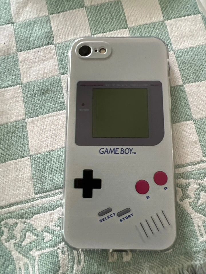 An iPhone fitted with the Gameboy Case For iPhone, featuring a retro Game Boy-themed silicone design that includes a screen, directional pad, and buttons marked "B," "A," "SELECT," and "START." The phone is placed on a checkered fabric surface.