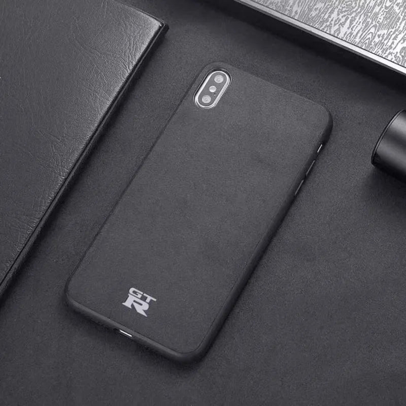 A GTR for iPhone, featuring a dual camera setup and encased in a sleek cover with the "Nissan GT-R" logo, is placed on a dark surface. Nearby are black objects including a notebook and what appears to be a pen.