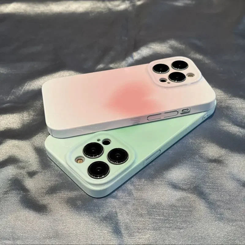 Two "Hearts for iPhone" smartphones with gradient-colored cases, one in pastel pink and the other in soft green, are placed on a shiny surface, the pink one atop. Both phones feature triple-lens cameras and stylish cases that enhance their gradient design.