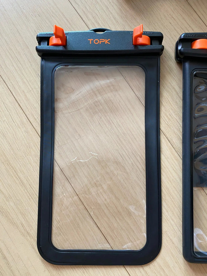 An IPX8 Waterproof Phone Pouch with a clear, waterproof design, featuring a black and orange frame labeled "TOPK," is lying on a wooden surface. This pouch, perfect for outdoor activities, includes a secure locking mechanism at the top.